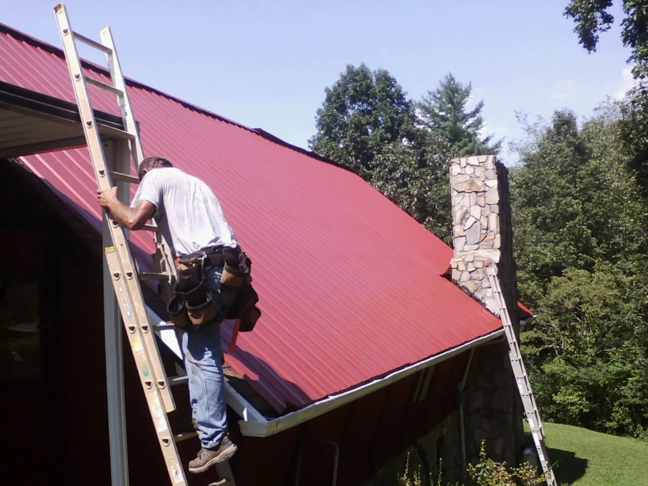 Roofer on a ladder beside a red metal roof and stone chimney — Soffit & Fascia Repair by Roofing Brothers in Kiamesha Lake